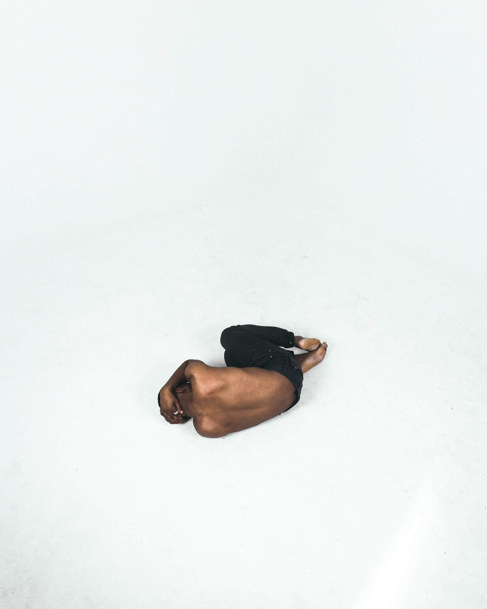 Foto afastada de um homem negro sem camisa deitado em posição fetal sobre um chão totalmente branco