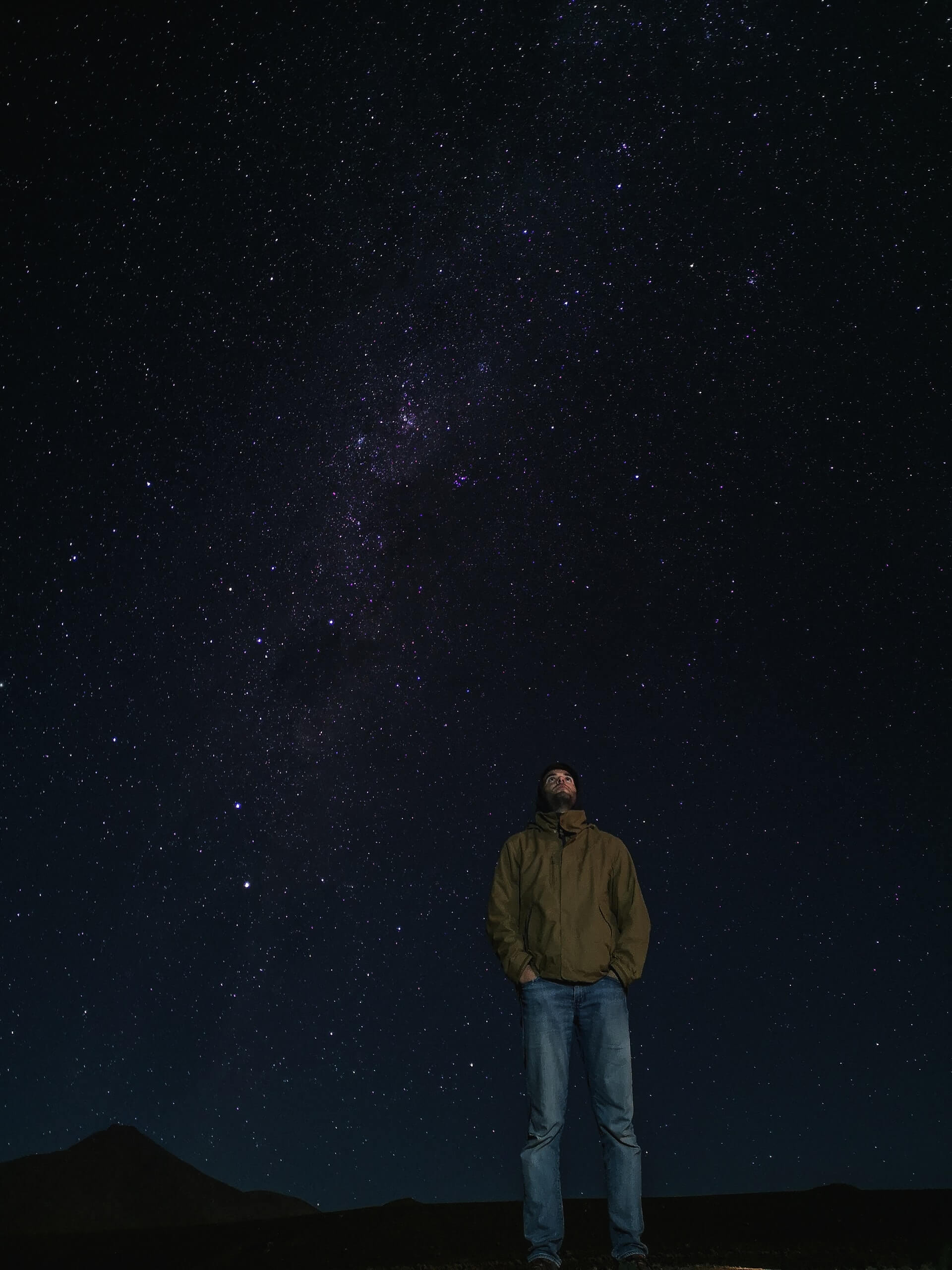 Foto de um homem de jaqueta marrom e calça jeans olhando para um céu bastante estrelado