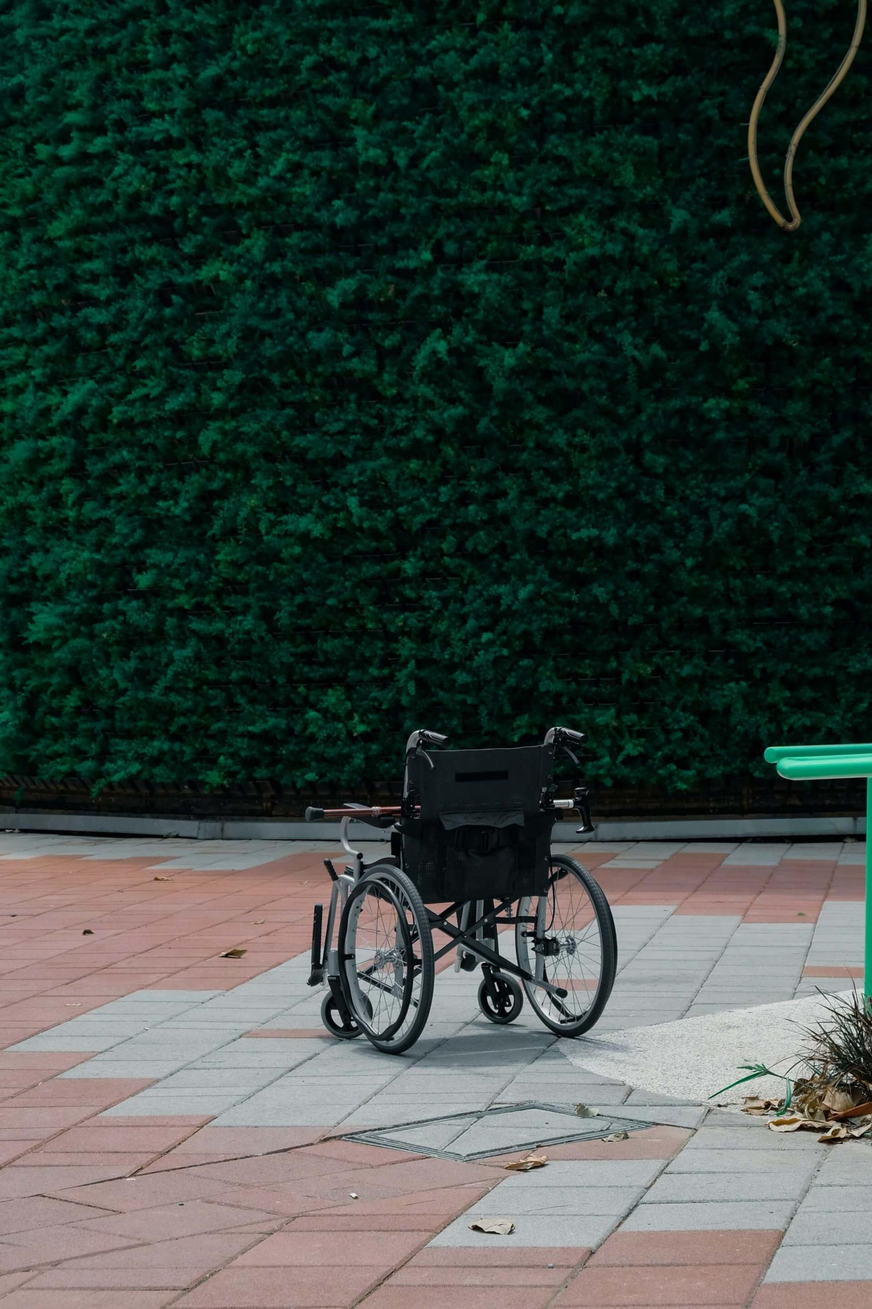 Foto de uma cadeira de rodas vazia com uma bengala sobre o assento, estacionada em um pátio com chão de placas de cimento alternadas entre cinza e avermelhado, em frente a uma cerca viva de plantas bem viçosas